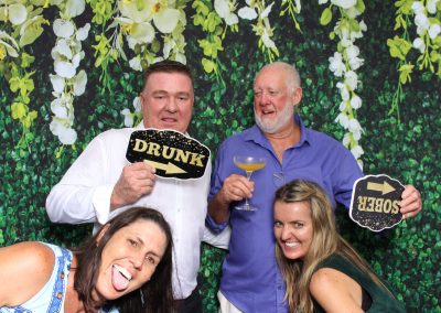Four people pose in front of a floral backdrop holding signs labeled "Drunk" and "Sober"; one person playfully sticks their tongue out.
