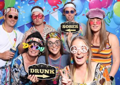 A group of seven people at a party wearing colorful, quirky costumes and novelty glasses. Two hold signs that read "DRUNK" and "SOBER