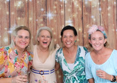 Four women in colorful attire posing in front of a wooden backdrop with string lights.