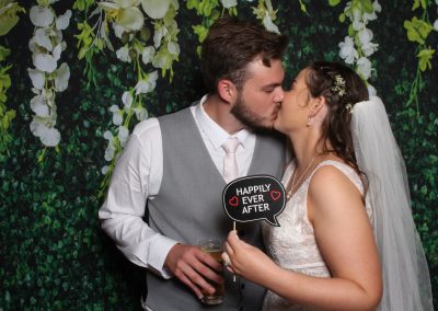 Couple at wedding holding a "Happily Ever After" sign, with greenery backdrop.