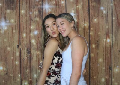 Two women in front of a wooden backdrop with twinkling lights, one in a floral dress, the other in white.